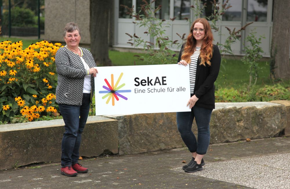 Zwei Personen halten ein Schild mit dem Logo der SekAE
